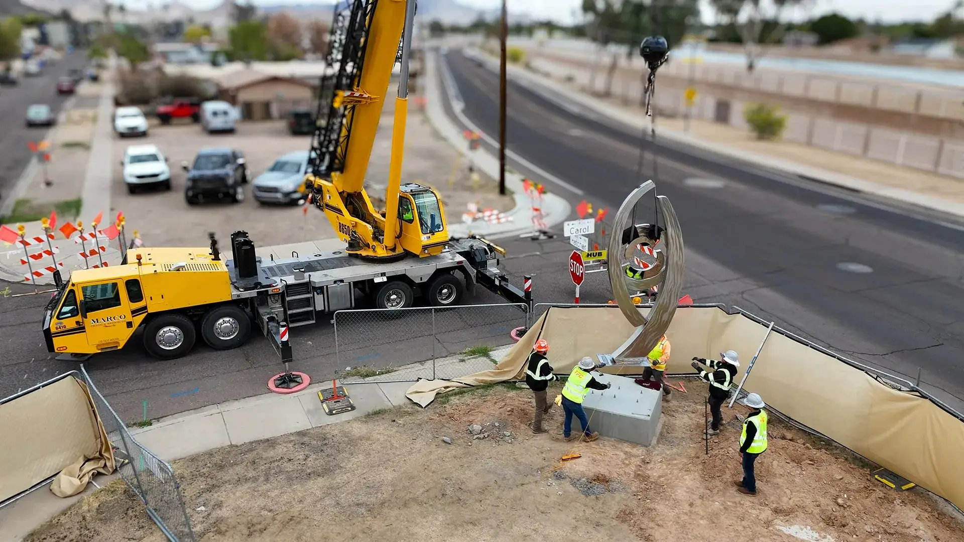 Crane lifting a large sculpture into place on a city street while crews watch