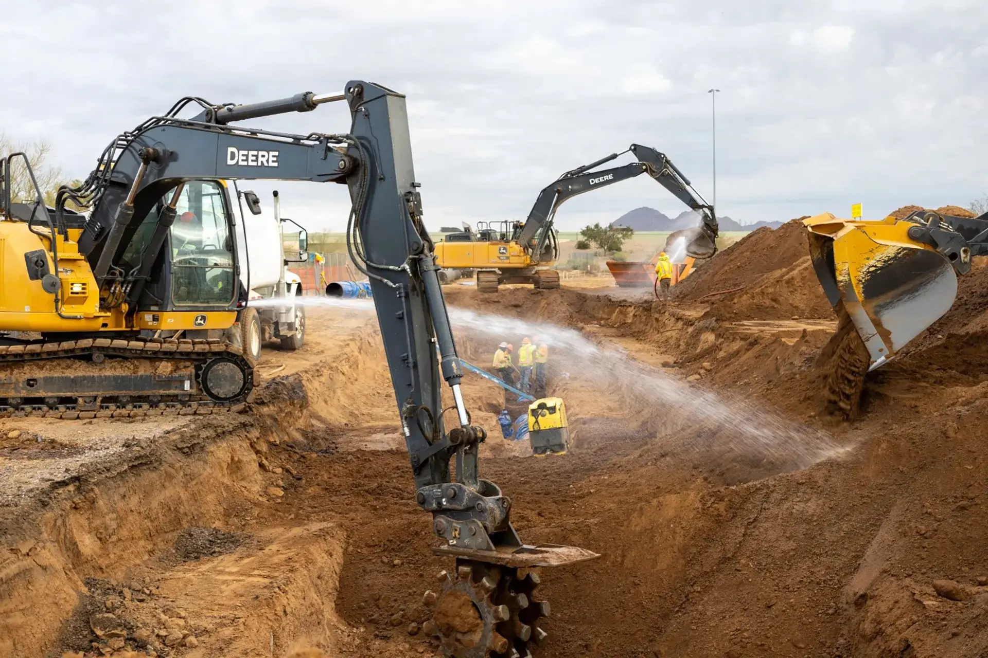 Excavator digging a trench while a worker sprays water to control dust